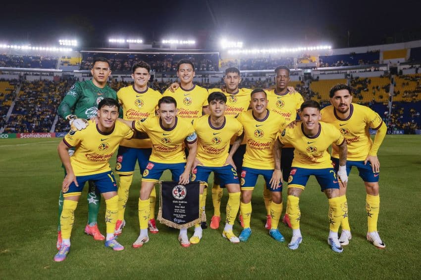 The Club America soccer team from Mexico City posing as a group in two horizontal rows some standing, some crouching slightly. They are all wearing their uniform yellow tee shirts and blue shorts. They are posing on the pitch of a stadium